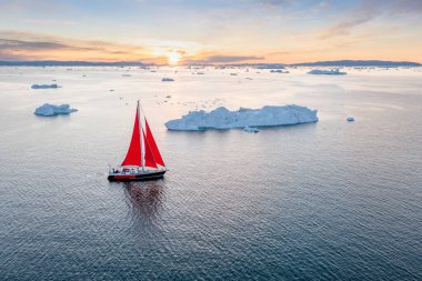 Sail boat with red sails cruising among ice bergs during dusk in front of a full moon. Disko Bay, Greenland. Midnight sun, romantic view. Climate change and global warming