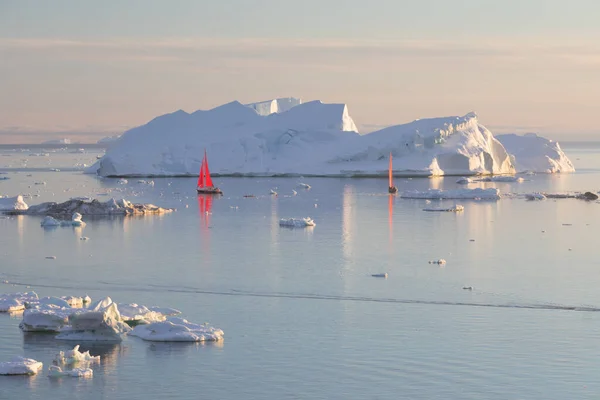 Sail boats with red sails cruising among ice bergs during dusk in front of a full moon. Disko Bay, Greenland. Midnight sun, romantic view. Climate change and global warming