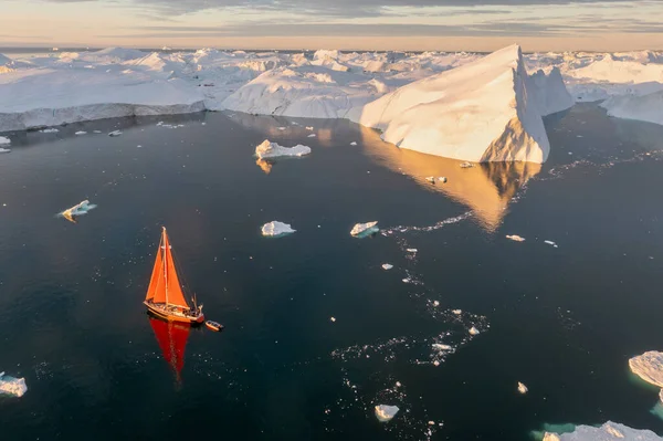 Sail boat with red sails cruising among ice bergs during dusk in front of a full moon. Disko Bay, Greenland. Midnight sun, romantic view. Climate change and global warming