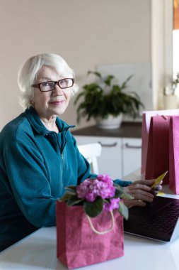 Çekici, gri saçlı 85 yaşında bir kadın internetten alışveriş yapıyor. O, elinde bir kredi kartı tutuyor. Satış konsepti.