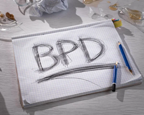 Concept of borderline personality disorder. The abbreviation for BPD is expressively written in pencil with strong pressure on paper. There is crumpled paper and a broken glass on the table. Strong emotional experience.