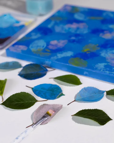 DIY Abstract botanical art. The picture is made by applying acrylic paints on the leaves and stamping on canvas. Blue on blue.
