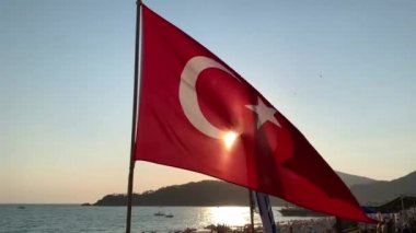 Bayrak denizin ve güneşin arka planında dalgalanıyor. Rüzgarda hilal şeklinde ay olan kırmızı Türk bayrağı. Türkiye, Oludeniz, Babadag Dağı, 8 Ağustos 2021