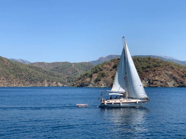 A yacht with a white sail on the open sea, against the backdrop of mountains. A boat with a sail on the waves of a calm sea. A sailboat floats in the ocean off the coast.