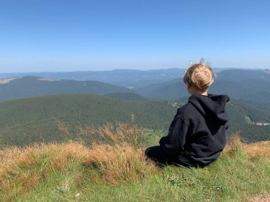 The boy is sitting on the top of the mountain. The child looks at the tops of the summer mountains. A man contemplates the ridges of the high Carpathians.