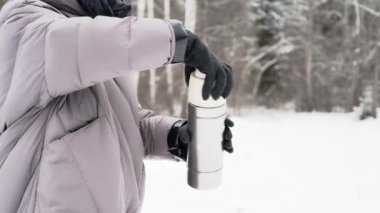 Videoda, siyah eldivenli kadın elleri termostan bir fincana sıcak kahve döküyor. Çam ağaçları, kar, arka planda kış.