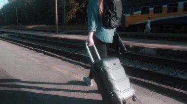 Elinde bavul ve siyah bir sırt çantası olan bir kadın turist tren platformu boyunca yavaşça yürüyor. Güneş parlıyor. Sağda hareket eden bir tren var..