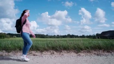 Ön planda siyah sırt çantalı bir turist kırsal yolda tek başına yürüyor. Kadın parlak güneşten kendini eliyle kapatıyor. Tarla, orman, gökyüzü ve bulutlar arka planda