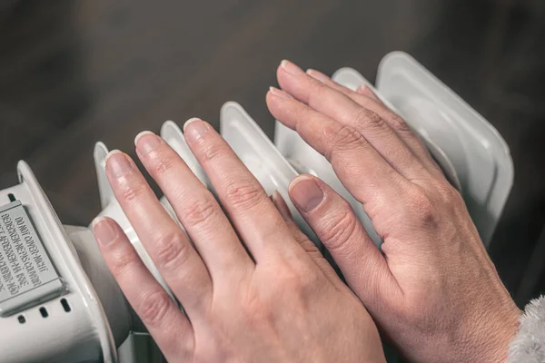 woman keeps hands warm over an oil-filled mobile heater. It\'s cold at home, electricity is expensive.