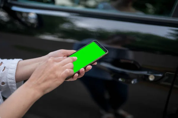 A woman holds a phone with a green screen in her hands against the background of a black car. a business woman stands with a phone by the car.