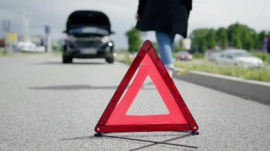 Red warning triangle and a woman walking towards her broken car on the road