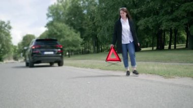 A woman walks along the side of the road and installs red triangular warning signal. Emergency stop sign on the road closeup. Blurred background