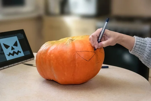A woman draws a scary face on a pumpkin with a black marker for Halloween. Festive decoration concept