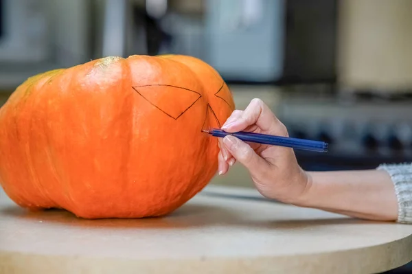 A woman draws a scary face on an orange pumpkin with a black marker in the kitchen. Halloween holiday design concept.