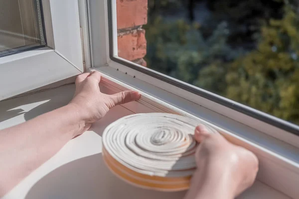 a woman glues a sealing rubber tape on a window in a living room.