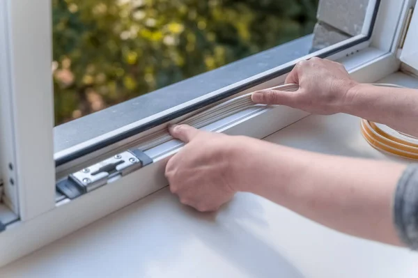 a woman glues a sealing rubber tape on a window in a living room.