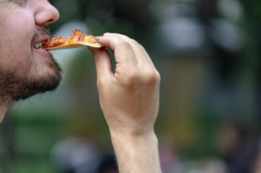 Man bites a slice of pizza. Close up caption. Outdoor background