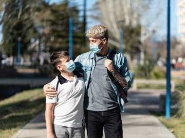 Caucasian father embracing his boy while walking to school on a sunny day wearing face masks. Back to school and virus spread concept