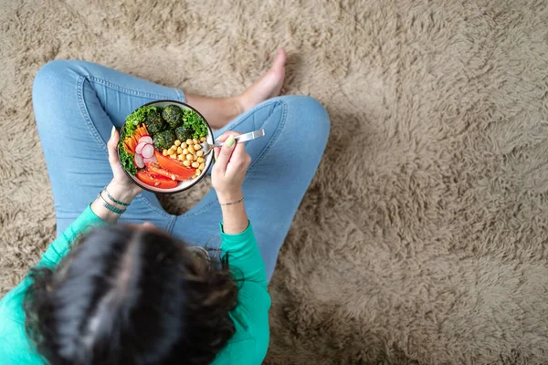Brunette woman eating veggie bowl in the floor of her living room. Copy space. Top view shot