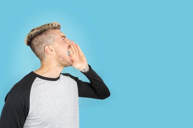 Açık mavi arka planda dikilen beyaz bir adam yukarı bakıyor ve birini çağırıyormuş gibi bağırıyor. İzole stüdyo çekimi. Reklamcılık için sağ tarafa boşluk kopyala