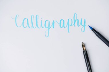 Calligrahpy kelimesi bir fırça kalem ve dolmakalemin yanında beyaz bir kağıda yazılmış, mavi keçeli kaleme odaklan.