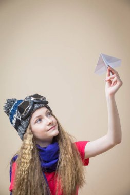 Uçan Gözlüklü Genç Kız ve Origami kağıt uçakla örülü kalın şapka pozu bej arka planda. Dikey Resim Yönlendirmesi