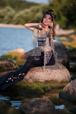 Deniz kıyısındaki Kayalıklarda Deniz Kızları 'nın özlemi. Deniz kabuğu desenli taç takıyor ve denizin kraliçesi gibi sert vücutla kaplı siyah parlak kuyruk takıyor. Dikey Resim Yönlendirmesi