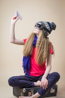 Uçan Dişliler içinde gülümseyen Kafkasyalı Genç Kız ve Sıcak Şapka Ören Origami Kağıt Uçağı Eski Bavul 'da Bej Arkaplan Üzerinde Otururken. Dikey Çekim