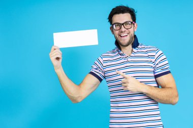 Mavi arkaplanda parmak hareketleriyle poz verirken elinde beyaz tabakla gülümseyen beyaz tenli yakışıklı Brunet Adam. Yatay Görüntü