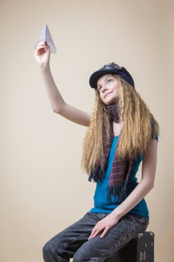 Uçan Gear 'lı Sarışın Gülümseyen Beyaz Kız, seyahat çantasının üzerinde otururken Origami Kağıt Uçağı' nı fırlatıyor. Dikey Resim Yönlendirmesi