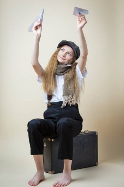 Beyaz üniformalı genç bir kız ve bej arka planda çıplak ayakla otururken Origami kağıt uçaklarıyla sokak şapkası pozu veriyor. Dikey Resim Yönlendirmesi