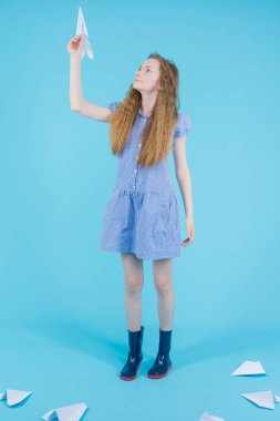 Gençlik yaşam tarzı. Blue Checked Dress ve Rubber Boots 'taki genç kız Origami Kağıt Uçağı' nı Mavi Arkaplan 'a fırlatıyor. Dikey Resim Yönlendirmesi