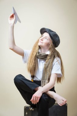 Okul üniformalı genç bir kız ve bej arka planda eski bir bavulda çıplak ayakla otururken Origami kâğıt uçakla sokak şapkası pozu veriyor. Yatay Resim