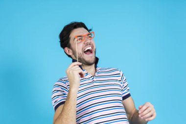 Komik, yakışıklı, beyaz bir adam sanatsal kağıt gözlüklü, kırmızı gözlüklü, kusursuz mavi arka planda poz veriyor. Yatay Resim Yönlendirmesi