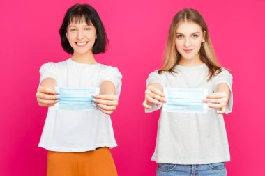 İki Olumlu Genç Kız Arkadaş, Pembe Arkaplana Karşı Yüz Koruma Maskesi Uygulaması Gösteriyor. Yatay Resim