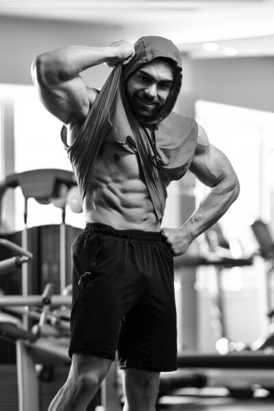 Healthy Young Man Standing Strong in the Gym and Flexing Muscles in a Hoodie - Muscular Athletic Bodybuilder Fitness Model Posing After Exercises