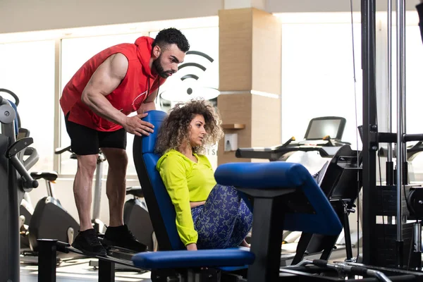 Young Adult Woman Working Out In Gym - Doing Legs Exercise On Machine With Help Of Her Personal Trainer