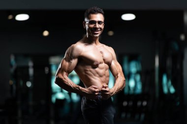 Gynecomastia göğüsleri olan genç adam spor salonunda güçlü duruyor ve kaslarını kasıyor.