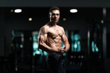 Gynecomastia göğüsleri olan yakışıklı genç adam spor salonunda güçlü duruyor ve kaslarını kasıyor.