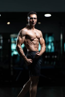 Gynecomastia göğüsleri olan yakışıklı genç adam spor salonunda güçlü duruyor ve kaslarını kasıyor.