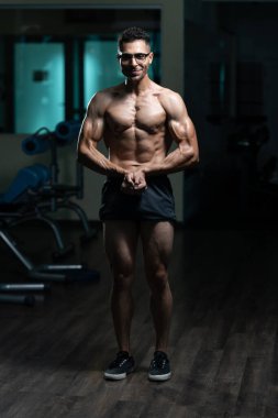 Gynecomastia göğüsleri olan genç adam spor salonunda güçlü duruyor ve kaslarını kasıyor.