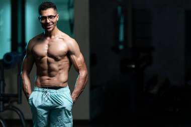 Gynecomastia göğüsleri olan genç adam spor salonunda güçlü duruyor ve kaslarını kasıyor.