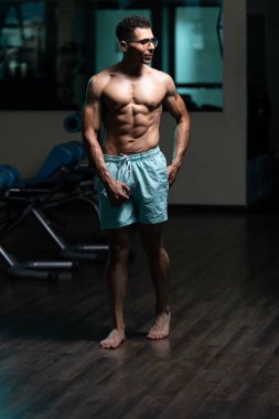 Gynecomastia göğüsleri olan genç adam spor salonunda güçlü duruyor ve kaslarını kasıyor.