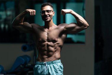 Gynecomastia göğüsleri olan genç adam spor salonunda güçlü duruyor ve kaslarını kasıyor.