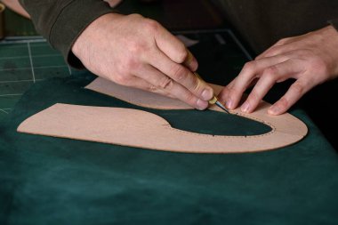 Shoemaker hands holding craft knife cutting green suede leather for handmade shoes on his workplace in workshop. Closeup. Copy space