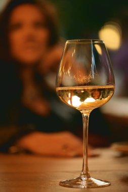 Kafede ya da restoranda tanınmayan bir kadının arka planında duran bir bardak beyaz şarap. Şarap kadehine odaklan. Yalnızlık ve rahatlama kavramı