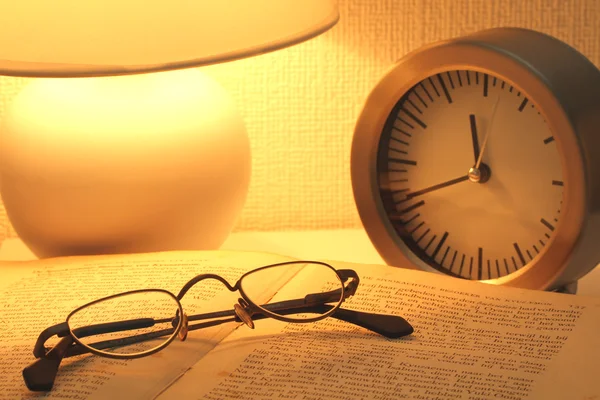 Brille auf dem Buch neben dem Wecker im Licht eines Nachtlichts — Stockfoto