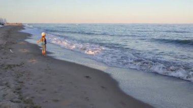 A lonely girl in a knitted hat and blue pants runs along a deserted sea beach with foamy waves lit by the setting sun