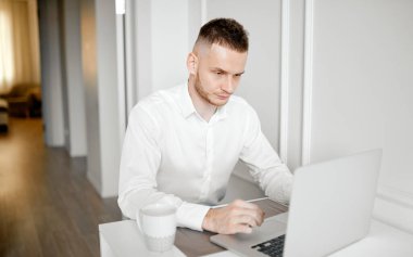 Müdür evde, mutfakta beyaz gömlekli ve fincanlı bir dizüstü bilgisayarın arkasında çalışıyor.