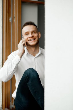 Mutlu adam pencere eşiğinde oturuyor ve telefonla konuşuyor.